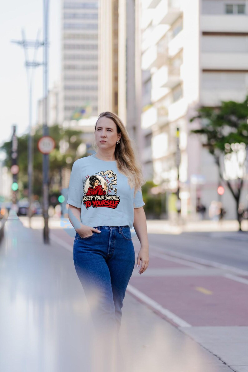 May include: A light blue short-sleeve t-shirt featuring a graphic illustration of a person in a red hoodie holding a cigarette with smoke swirling around. The text "KEEP YOUR SMOKE TO YOURSELF" is printed in bold red and black lettering below the figure.