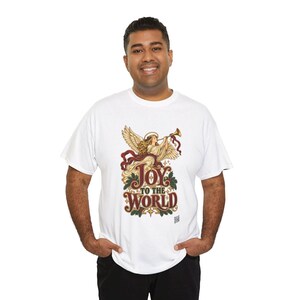 May include: A white short-sleeved t-shirt featuring a graphic of an angel playing a golden trumpet. Below the angel, the text reads Joy to the World in a decorative font, surrounded by green holly leaves and red ribbons on a white background.