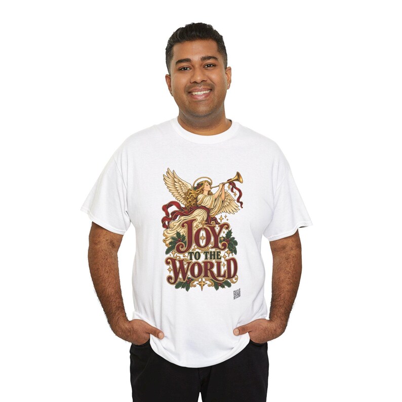 May include: A white short-sleeved t-shirt featuring a graphic of an angel playing a golden trumpet. Below the angel, the text reads Joy to the World in a decorative font, surrounded by green holly leaves and red ribbons on a white background.