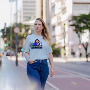 May include: A light blue short-sleeved t-shirt featuring a graphic print of a person in a blue shirt with the text ASK ME ANYTHING, along with smaller icons and decorative elements, paired with dark blue denim jeans.