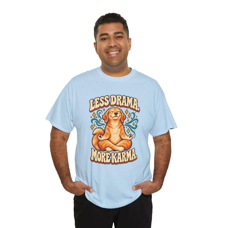 May include: A light blue short-sleeve t-shirt featuring a graphic of a golden dog sitting in a lotus meditation pose. The text above the dog reads LESS DRAMA and the text below reads MORE KARMA in a retro, rounded, multi-colored font.