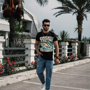 May include: A black short-sleeve t-shirt featuring a graphic of a fish jumping through waves with the text "CURRENTLY FIGHTING THE CURRENT" in yellow and white lettering, paired with blue denim jeans and white athletic sneakers.