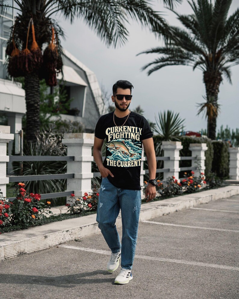 May include: A black short-sleeve t-shirt featuring a graphic of a fish jumping through waves with the text "CURRENTLY FIGHTING THE CURRENT" in yellow and white lettering, paired with blue denim jeans and white athletic sneakers.