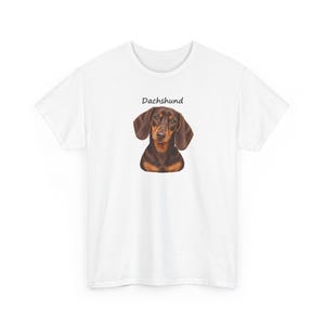 K&ouml;nnte beinhalten: Ein wei&szlig;es Kurzarm-T-Shirt mit einer zentralen Illustration eines braun-schwarzen Dackels. &Uuml;ber dem Kopf des Hundes ist das Wort Dachshund in einer einfachen schwarzen serifenlosen Schrift gedruckt. Das T-Shirt hat einen Rundhalsausschnitt.