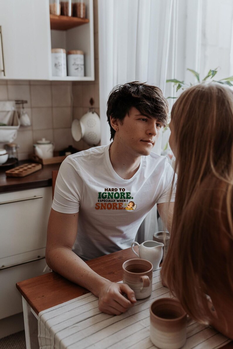 May include: A white short-sleeved t-shirt featuring the text "HARD TO IGNORE. ESPECIALLY WHEN I SNORE." in green and orange lettering, accompanied by a small graphic of a person sleeping in a bed.