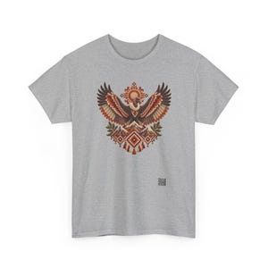 May include: A heather gray t-shirt featuring a central graphic of a large bird with wings spread wide above a mountain range. The design incorporates geometric patterns in shades of brown, orange, and cream, with a small QR code at the bottom.
