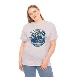 May include: A light gray short-sleeve t-shirt featuring a graphic print of a pilot and a small robot in a cockpit. The text reads WATCH OUT! I'M ON AUTOPILOT NOW. The design is circular with blue and white accents and a small airplane icon.