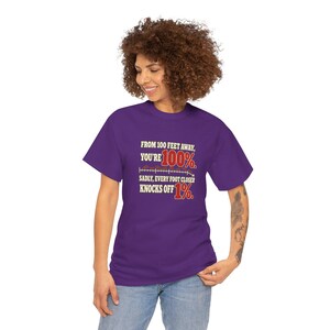 May include: A purple short-sleeve t-shirt featuring the text: FROM 100 FEET AWAY, YOU'RE 100%. SADLY, EVERY FOOT CLOSER KNOCKS OFF 1%. The text is printed in a mix of cream and red lettering with a graphic of a downward sloping line.
