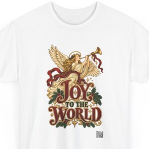 May include: A white t-shirt featuring a graphic illustration of an angel with blonde hair and wings playing a golden trumpet. Below the angel, the text reads JOY TO THE WORLD in large, decorative red and gold lettering, accented by green holly leaves.