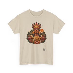 May include: A beige short-sleeved t-shirt featuring a central graphic of a person wearing a feathered headdress and ornate gold jewelry. The design includes mountain silhouettes, a sun, and geometric patterns in red, green, and gold.