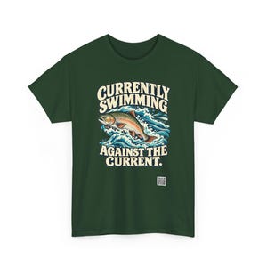 May include: A dark green t-shirt featuring a graphic of a trout jumping through blue waves. The text above the fish reads "CURRENTLY SWIMMING" and the text below reads "AGAINST THE CURRENT." A small square QR code is printed near the bottom.