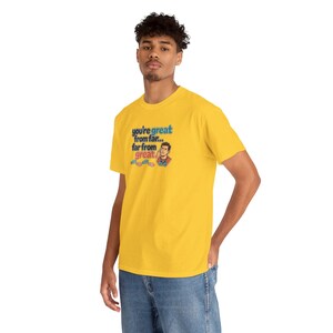 May include: A bright yellow short-sleeve t-shirt featuring a graphic print with the text 'you're great from far... far from great.' The design includes a small illustration of a person pointing to their head and three small game controllers.