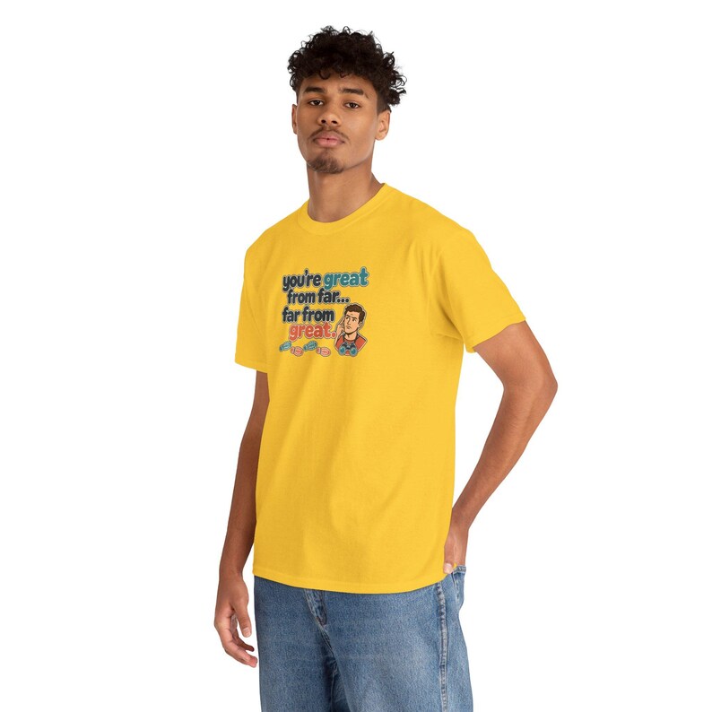 May include: A bright yellow short-sleeve t-shirt featuring a graphic print with the text 'you're great from far... far from great.' The design includes a small illustration of a person pointing to their head and three small game controllers.
