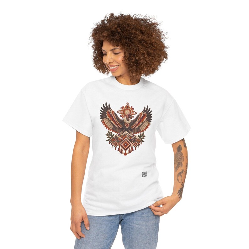 May include: A white short-sleeve t-shirt featuring a central graphic of a stylized condor with wings spread wide, positioned above mountain peaks and geometric patterns in shades of brown, orange, and cream, with a small QR code near the hem.