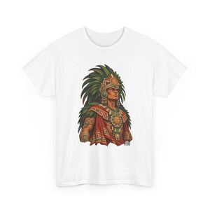 May include: A white short-sleeve t-shirt featuring a graphic print of a figure wearing a large green feathered headdress, a gold sun-disk chest piece, and a red cloak with geometric patterns. The figure has dark hair and facial markings.