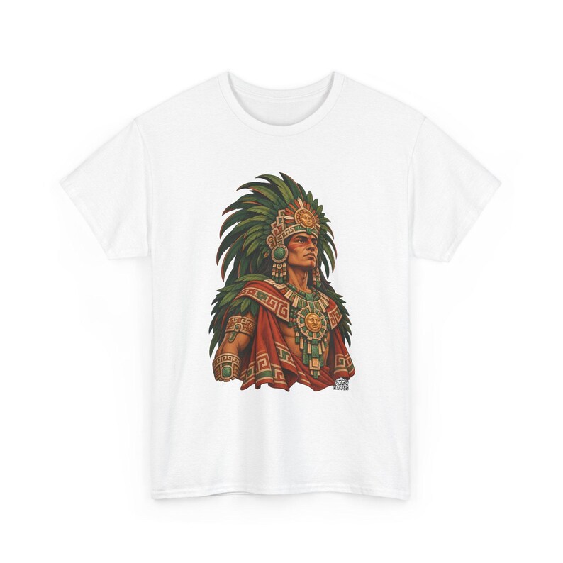 May include: A white short-sleeve t-shirt featuring a graphic print of a figure wearing a large green feathered headdress, a gold sun-disk chest piece, and a red cloak with geometric patterns. The figure has dark hair and facial markings.