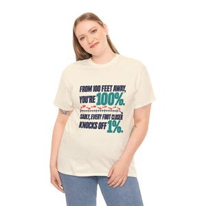 May include: A cream-colored short-sleeve t-shirt featuring dark blue and teal text that reads: FROM 100 FEET AWAY, YOU'RE 100%. SADLY, EVERY FOOT CLOSER KNOCKS OFF 1%. A graphic of a measuring line with small orange footprints is included.