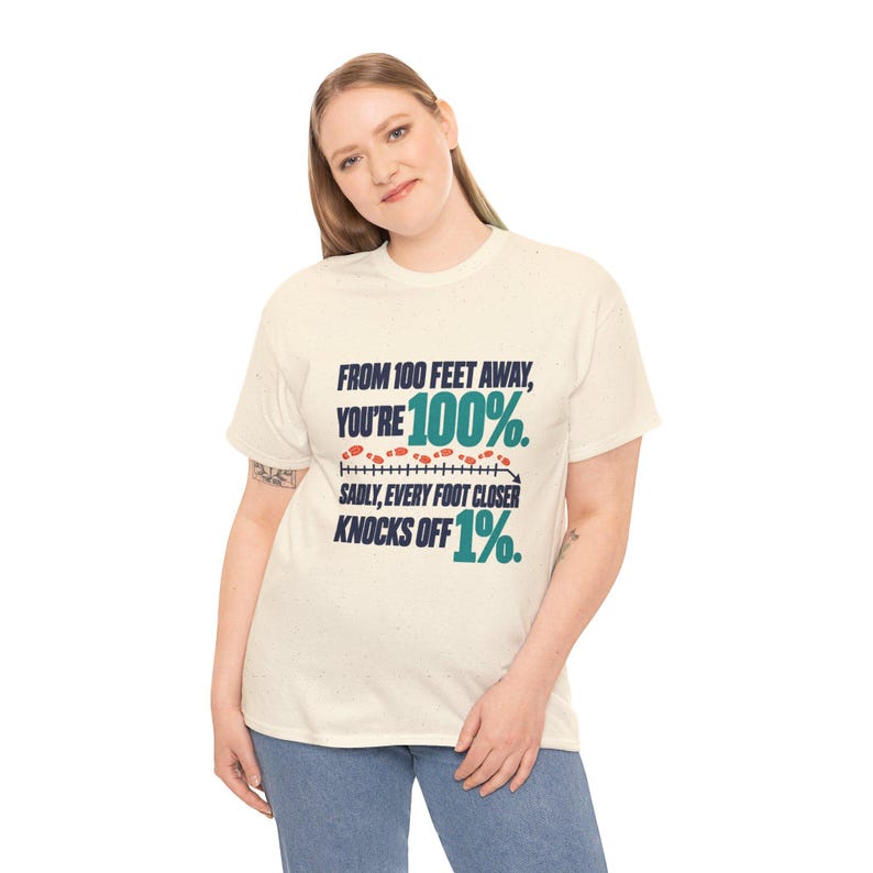 May include: A cream-colored short-sleeve t-shirt featuring dark blue and teal text that reads: FROM 100 FEET AWAY, YOU'RE 100%. SADLY, EVERY FOOT CLOSER KNOCKS OFF 1%. A graphic of a measuring line with small orange footprints is included.