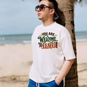 May include: A white short-sleeved t-shirt featuring the text "YOU ARE WELCOME... TO LEAVE!" in dark green and brown lettering, accompanied by a small brown door graphic, paired with bright blue drawstring shorts and dark sunglasses.