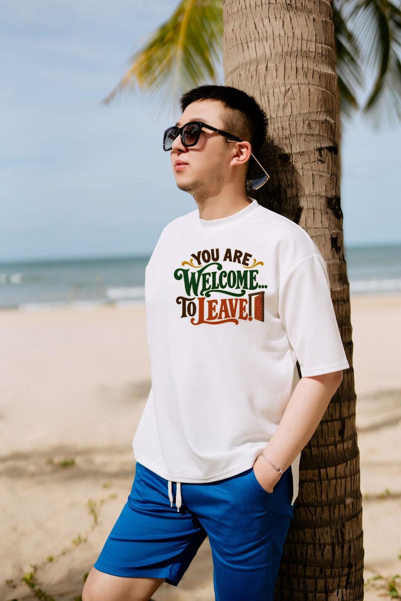 May include: A white short-sleeved t-shirt featuring the text "YOU ARE WELCOME... TO LEAVE!" in dark green and brown lettering, accompanied by a small brown door graphic, paired with bright blue drawstring shorts and dark sunglasses.