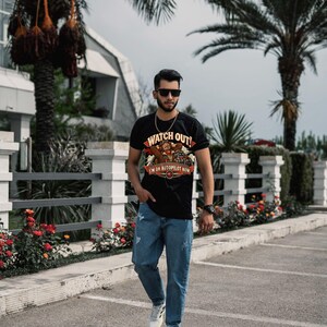May include: A black short-sleeved t-shirt featuring a graphic print of a pilot with the text "WATCH OUT! I'M ON AUTOPILOT NOW" in red and yellow, paired with light blue distressed denim jeans and white athletic sneakers.