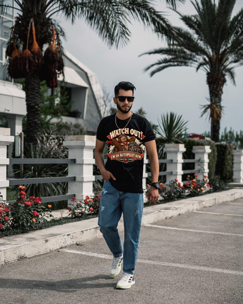 May include: A black short-sleeved t-shirt featuring a graphic print of a pilot with the text "WATCH OUT! I'M ON AUTOPILOT NOW" in red and yellow, paired with light blue distressed denim jeans and white athletic sneakers.