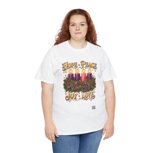 May include: A white t-shirt featuring a graphic of an Advent wreath with four lit candles, three purple and one pink, surrounded by greenery and ribbons. The text "HOPE PEACE" appears above the candles, and "JOY LOVE" appears below in gold.