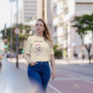 May include: A cream-colored short-sleeve t-shirt featuring a graphic of a white poodle with a pink bow on its head. Above the poodle, the text reads Poodle's Day in a simple black font. The shirt has a crew neckline and a straight hem.