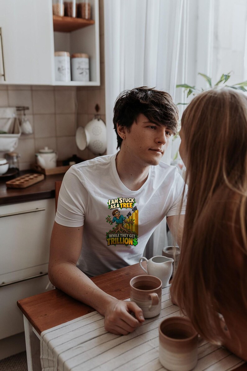 May include: A white short-sleeved t-shirt featuring a graphic print of a person with tree roots for legs. The text reads, "I AM STUCK AS A TREE WHILE THEY GET TRILLIONS," with the word "TRILLIONS" in green and a yellow bar graph background.