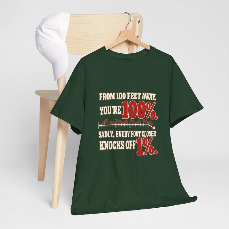 May include: A dark green t-shirt featuring the text "FROM 100 FEET AWAY, YOU'RE 100%. SADLY, EVERY FOOT CLOSER KNOCKS OFF 1%." in white and red lettering, with a small graphic of a downward trending line and red marks.