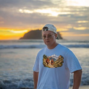 Puede incluir: Una camiseta blanca de manga corta con un estampado gr&aacute;fico rectangular de dos figuras caminando con una llama a trav&eacute;s de un paisaje monta&ntilde;oso con picos nevados, usada con una gorra de b&eacute;isbol de color claro y gafas.