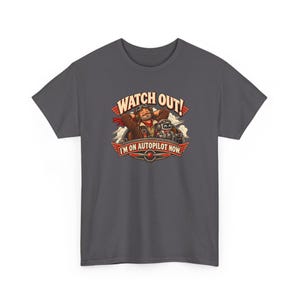 May include: A charcoal gray t-shirt featuring a graphic of a pilot with hands behind their head and a small robot in a cockpit. The text reads WATCH OUT! I'M ON AUTOPILOT NOW. The design includes red wings and a propeller emblem at the base.