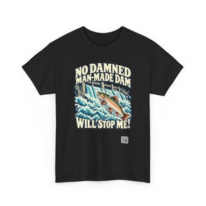 Puede incluir: Una camiseta negra con una ilustraci&oacute;n gr&aacute;fica de un pez saltando sobre una presa con agua blanca salpicando. El texto NO DAMNED MAN-MADE DAM WILL STOP ME! aparece en color crema. Un peque&ntilde;o c&oacute;digo QR est&aacute; impreso debajo del gr&aacute;fico.