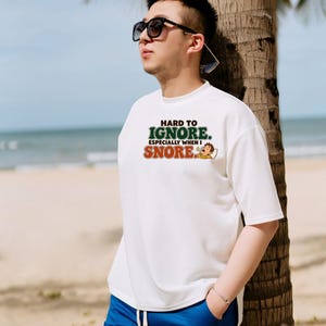 May include: A white short-sleeved t-shirt featuring the text "HARD TO IGNORE. ESPECIALLY WHEN I SNORE." in green and orange lettering, accompanied by a small graphic of a person sleeping with Zs floating above their head.