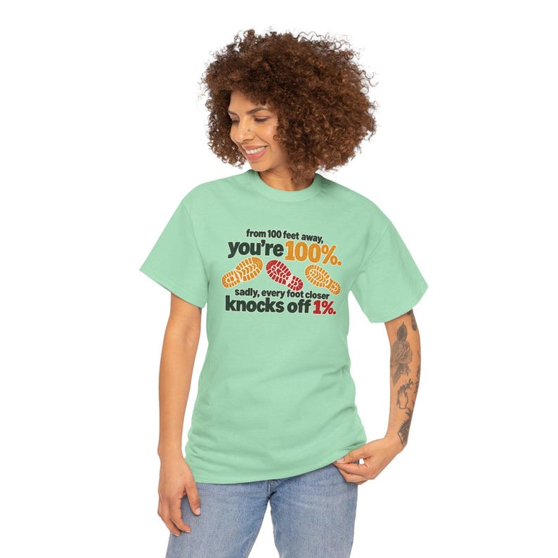 May include: A light green short-sleeve t-shirt featuring black text that reads from 100 feet away, you're 100%. sadly, every foot closer knocks off 1%. The design includes three orange and red shoe print graphics across the chest area.