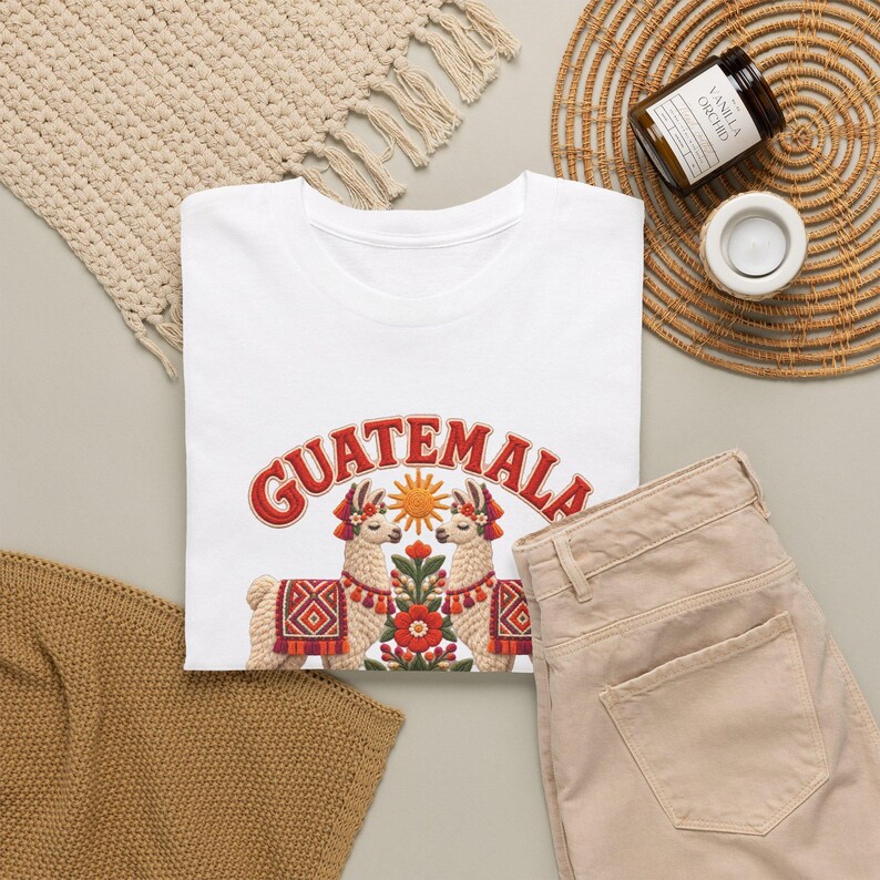 Puede incluir: Una camiseta blanca con un estampado gr&aacute;fico que muestra la palabra GUATEMALA en letras rojas sobre dos llamas frente a frente. Entre las llamas hay un sol estilizado y un arreglo floral en tonos rojos, naranjas y verdes.