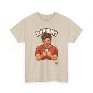 May include: A beige short-sleeved t-shirt featuring a graphic illustration of a person in a red shirt looking down at a smartphone. Above the figure, the text reads R U PHONY? in bold, cream-colored letters with a dark outline.