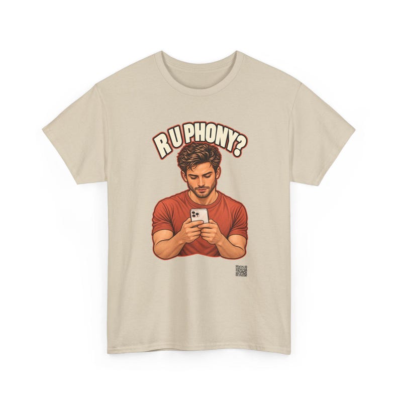 May include: A beige short-sleeved t-shirt featuring a graphic illustration of a person in a red shirt looking down at a smartphone. Above the figure, the text reads R U PHONY? in bold, cream-colored letters with a dark outline.