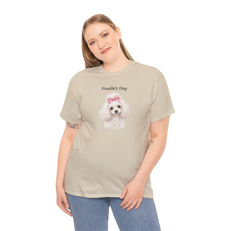 May include: A beige short-sleeved t-shirt featuring a graphic of a white poodle with a pink bow on its head. Above the poodle, the text reads Poodle's Day in a simple black font. The shirt has a crew neckline and a standard fit.