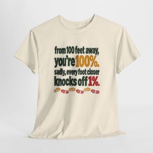 May include: A cream-colored short-sleeve t-shirt featuring the black text: from 100 feet away, you're 100%. sadly, every foot closer knocks off 1%. The numbers 100% and 1% are highlighted in orange and red, with small colorful footprints below.