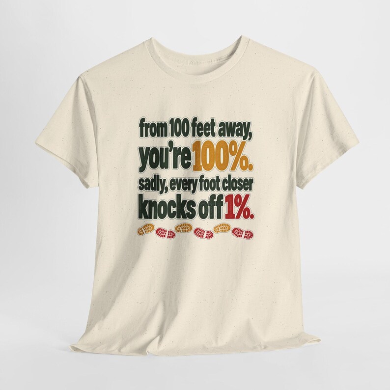 May include: A cream-colored short-sleeve t-shirt featuring the black text: from 100 feet away, you're 100%. sadly, every foot closer knocks off 1%. The numbers 100% and 1% are highlighted in orange and red, with small colorful footprints below.