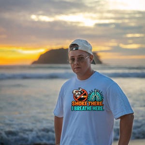 May include: A white short-sleeved t-shirt featuring a graphic print of a no-smoking symbol next to a pair of blue lungs. The text reads, "SMOKE THERE. I BREATHE HERE." in orange and teal lettering centered on the chest of the garment.