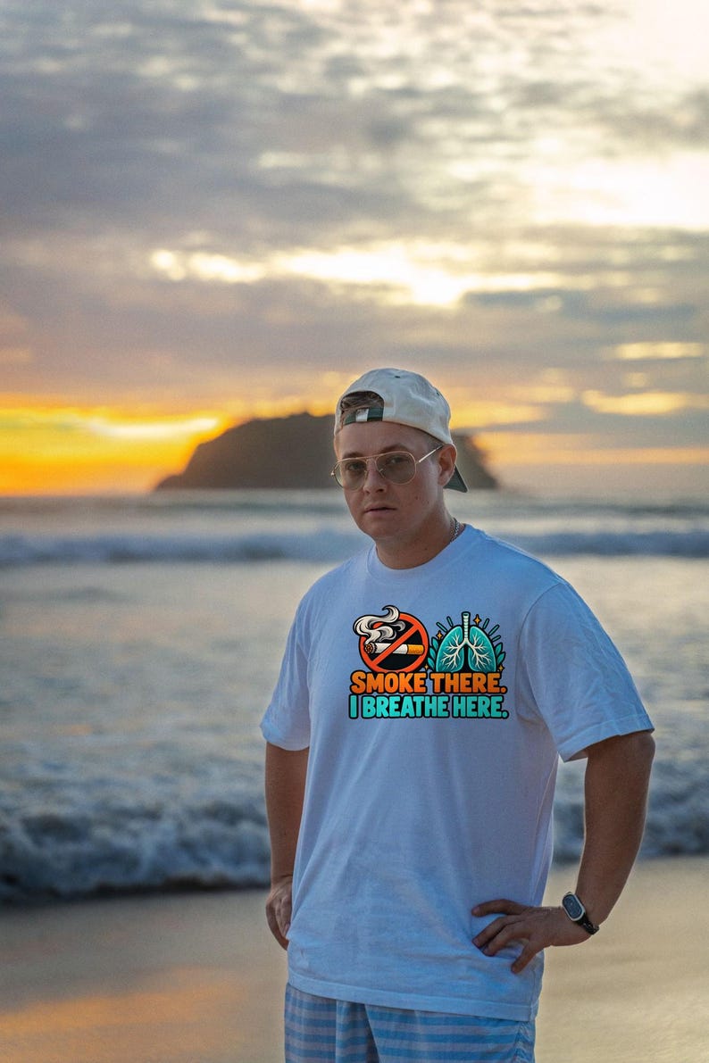 May include: A white short-sleeved t-shirt featuring a graphic print of a no-smoking symbol next to a pair of blue lungs. The text reads, "SMOKE THERE. I BREATHE HERE." in orange and teal lettering centered on the chest of the garment.