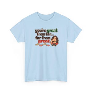 May include: A light blue t-shirt featuring a graphic with the text "you're great from far... far from great." The design includes a cartoon illustration of a person looking through binoculars and a series of small colorful footprints.