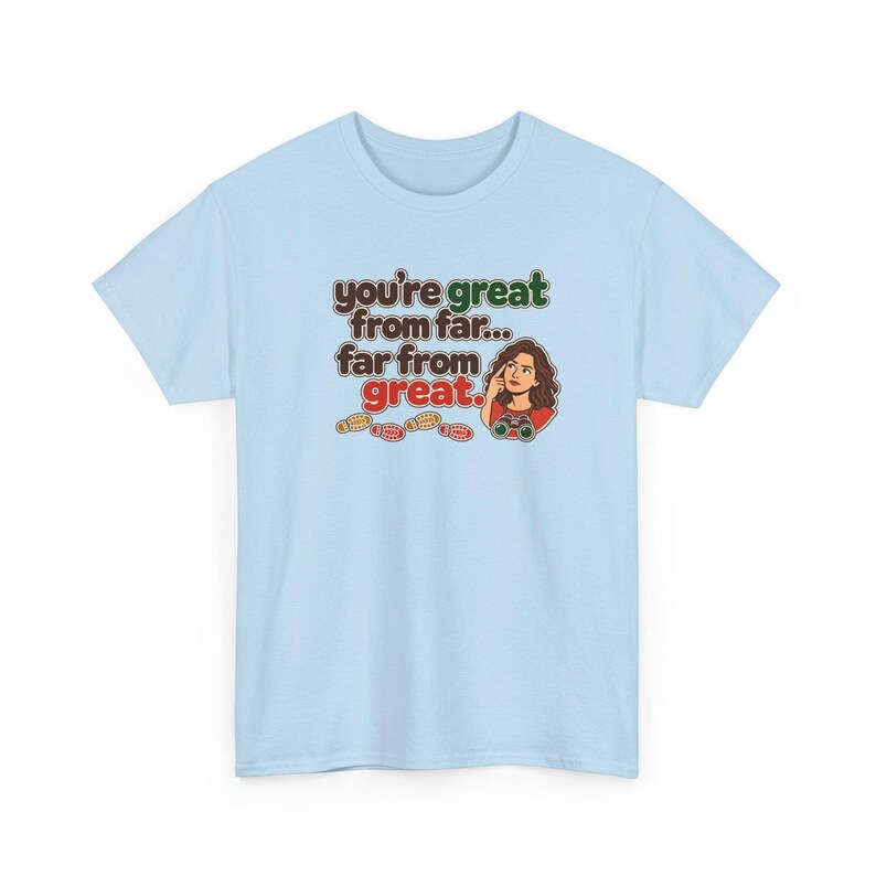 May include: A light blue t-shirt featuring a graphic with the text "you're great from far... far from great." The design includes a cartoon illustration of a person looking through binoculars and a series of small colorful footprints.