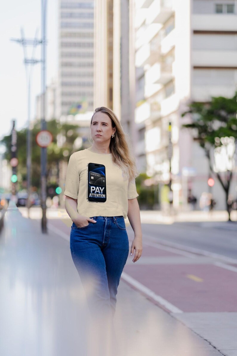 May include: A cream-colored short-sleeve t-shirt featuring a graphic of a smartphone displaying a credit card and the text PAY ATTENTION. The shirt is paired with blue denim jeans, creating a casual outfit suitable for daily wear.