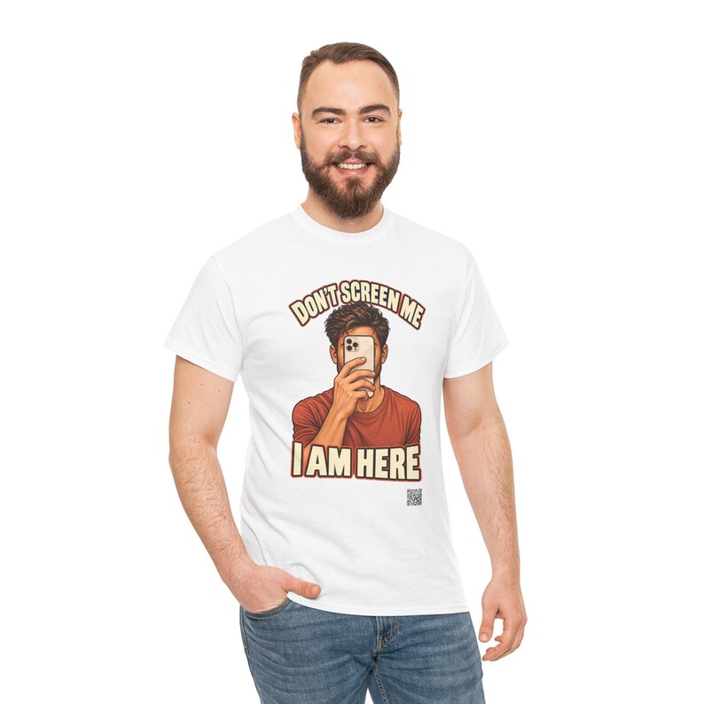 Op de afbeelding: Een wit T-shirt met korte mouwen en een afbeelding van een persoon in een roestkleurig shirt die een smartphone voor het gezicht houdt. De tekst boven en onder de afbeelding luidt "DON'T SCREEN ME I AM HERE" in dikke, beige hoofdletters.
