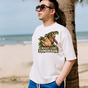 Pode incluir: Uma t-shirt branca de manga curta com um gr&aacute;fico de duas pregui&ccedil;as castanhas penduradas em ramos de &aacute;rvore verdes. Abaixo das pregui&ccedil;as, o texto I MIGHT COME TOMORROW est&aacute; escrito em letras claras e negrito com contorno escuro.