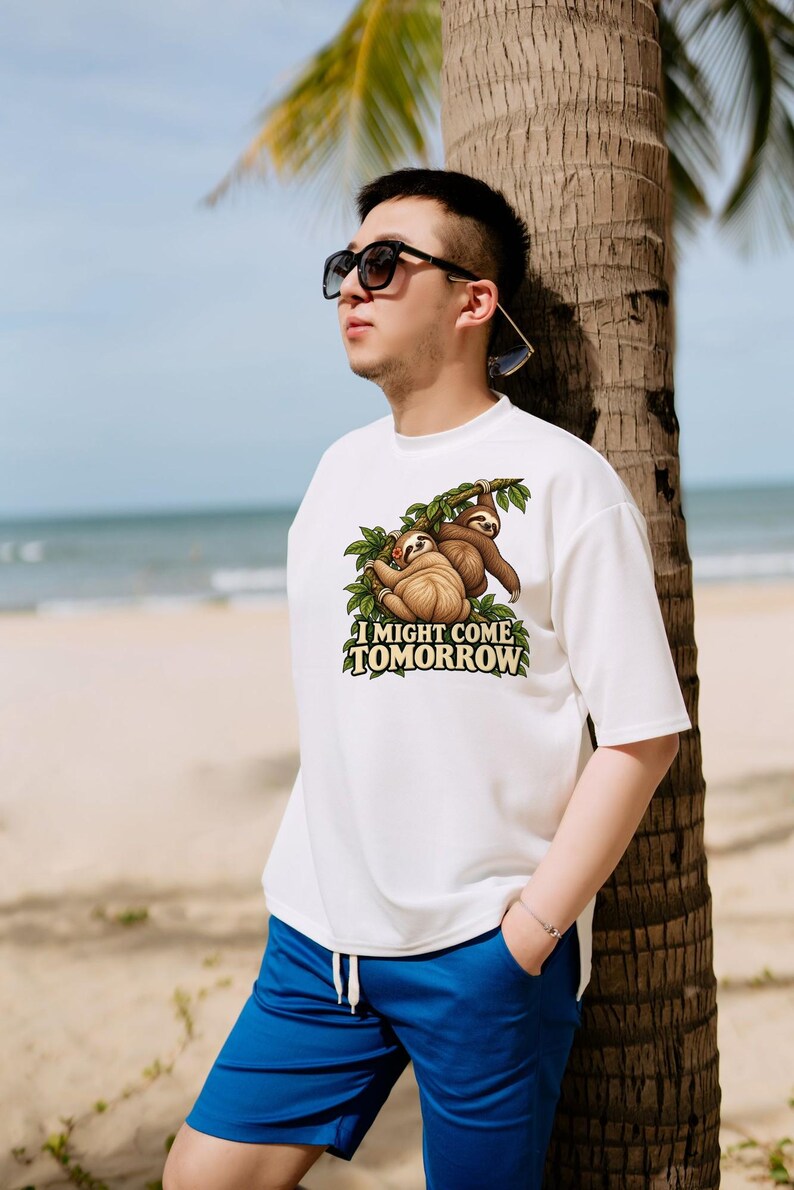 Pode incluir: Uma t-shirt branca de manga curta com um gr&aacute;fico de duas pregui&ccedil;as castanhas penduradas em ramos de &aacute;rvore verdes. Abaixo das pregui&ccedil;as, o texto I MIGHT COME TOMORROW est&aacute; escrito em letras claras e negrito com contorno escuro.