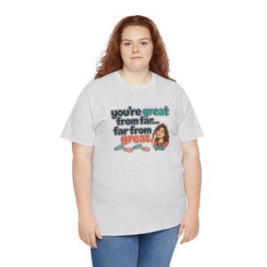 May include: A light gray short-sleeve t-shirt featuring a graphic print with the text "you're great from far... far from great." The design includes a cartoon illustration of a person looking through binoculars and several small footprint icons.